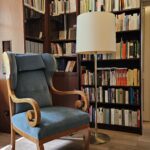 Das Bild zeigt die Bibliothek des Bonhoeffer-Hauses mit deckenhohen Bücherregalen, einem großen Sessel und einer Stehlampe. The picture shows the library of the Bonhoeffer House with ceiling-high bookshelves, a large armchair and a floor lamp.