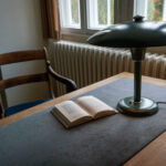 Das Bild zeigt Dietrich Bonhoeffers historischen Schreibtisch am Fenster seines ehemaligen Studierzimmers. Darauf befinden sich ein Buch und eine Schreibtischlampe.