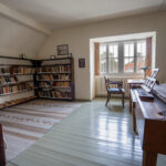 Das Bild zeigt das ehemalige Studierzimmer Dietrich Bonhoeffers im rekonstruierten Zustand von 1943: grüner Holzboden und braune Holzmöbel, links Bücherregale und ein Bett, rechts ein Klavikord sowie am Fenster ein Schreibtisch mit Stuhl. The picture shows Dietrich Bonhoeffer's former study room as reconstructed in 1943: green wooden floor and brown wooden furniture, bookshelves and a bed on the left, a clavicle on the right and a desk and chair by the window.