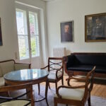Das Bild zeigt das Wohnzimmer des Bonhoeffer-Hauses mit Sofa, Tisch und Stühlen mit Familienproträts an den Wänden. The picture shows the living room of the Bonhoeffer House with two sofa, a table, armchairs and family portraits.